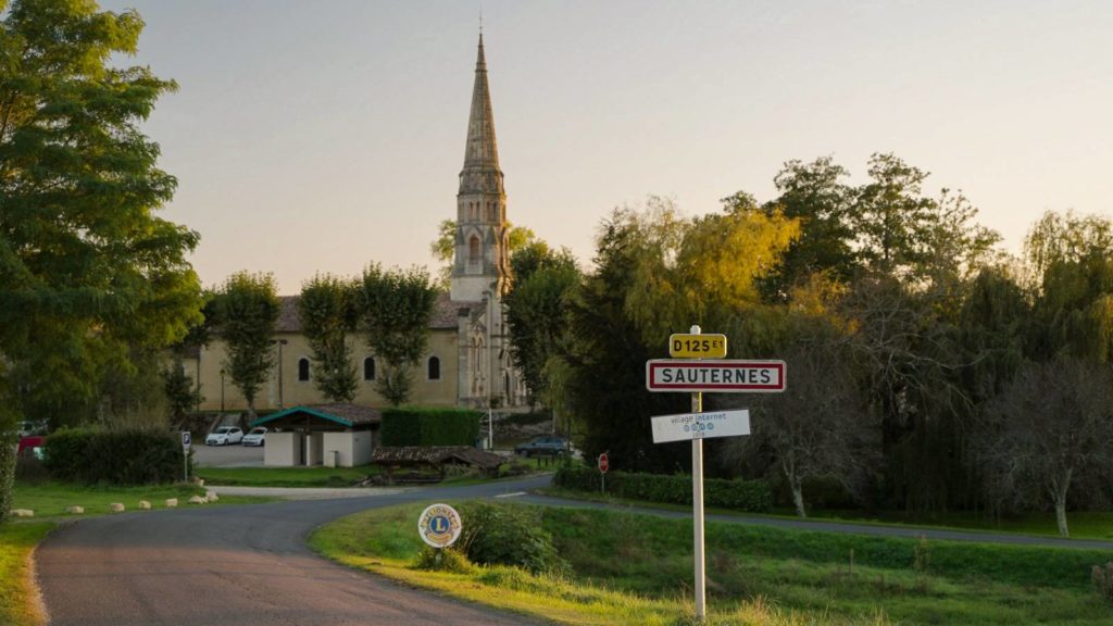 Entrance sign to the village of Sauternes with a historic church in the background, captured during a sunset on a Bordeaux wine tour.